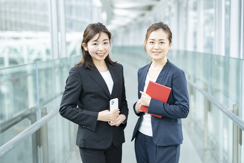 Two corporate insurance brokers in business suits smiling at the camera, holding a smartphone and a red folder, in a modern office walkway.