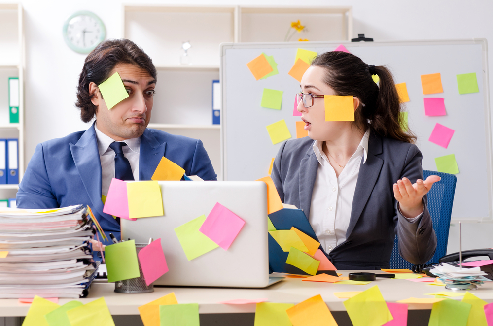 Two corporate insurance brokers at a desk covered in colorful sticky notes, one looking puzzled and the other speaking, in a cluttered workspace.