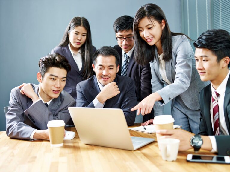 A group of six people in business attire gather around a laptop on a table, discussing an insurance plan. One woman points at the screen while others observe, making comparisons. Two cups of coffee are also on the table.