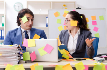 Two corporate insurance brokers at a desk covered in colorful sticky notes, one looking puzzled and the other speaking, in a cluttered workspace.