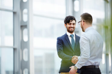 Two businessmen shaking hands in a modern office building discussing employee benefits in Singapore, with glass windows in the background.