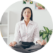 A woman in business attire sits cross-legged on a desk, meditating with eyes closed in a bright, modern home office.