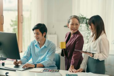 Three people in business attire collaborate in an office; one types at a computer, while the others stand nearby holding a mug and a tablet, discussing employee benefits in Singapore.